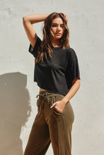 Load image into Gallery viewer, Woman wearing a black t-shirt and brown pants against a light gray wall.
