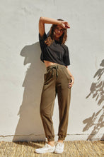 Load image into Gallery viewer, Woman wearing a black crop top and olive green pants against a white wall.
