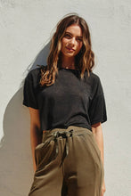 Load image into Gallery viewer, Woman wearing a black t-shirt and olive green pants against a light gray wall
