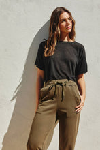 Load image into Gallery viewer, Woman wearing a black t-shirt and olive green pants against a gray wall
